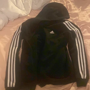 Svart hoodie från Adidas - Säljer en svart hoodie från Adidas med tre vita ränderna längs ärmarna. Den har en dragkedja framtill och är i ganska bra skick. Är lite nopprig på vissa ställen (se bild)
