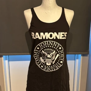 Svart Ramones linne - Säljer ett svart linne med Ramones-tryck. Linnet har ett stort vitt tryck med bandets namn och emblem på framsidan. Perfekt för sommarens festivaler eller en avslappnad dag. Passar både till jeans och shorts!