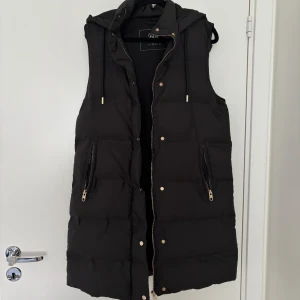 Svart dunväst från Zara - Säljer en snygg svart dunväst från Zara. Den har en huva, stängs med dragkedja och knappar. Perfekt för höst och vinter, och den är i nyskick! Den är i stl Xs för den är stor i storleken, passar S&M och går även att ha en tjock tröja under