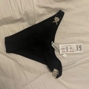 Svart bikiniunderdel  - Snygg svart bikiniunderdel från med dekorativa gulddetaljer på sidorna. Perfekt för en stilren look på stranden eller vid poolen. 🖤köpt i fel storlek, helt oanvända! Taggen fortfarande på!