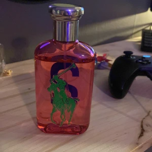 Röd parfymflaska från Ralph Lauren - Snygg röd parfymflaska från Ralph Lauren med en silverfärgad kork. Flaskan har en grön logotyp med en polospelare och en stor blå siffra 2 på framsidan. Perfekt för den som gillar en sportig och elegant stil. 
