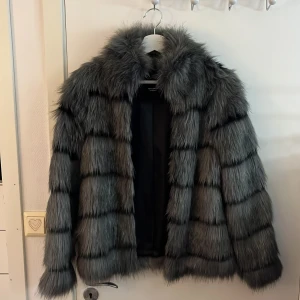 Grå pälsjacka - Snygg och fluffig grå pälsjacka med långa ärmar. Perfekt för att hålla sig varm och stilren under kyliga dagar. Jackan har en klassisk design med horisontella ränder och en mjuk krage.
