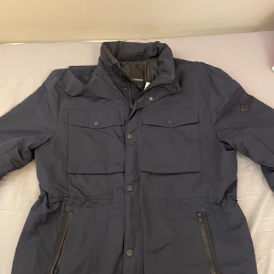 Mörkblå field jacket från J.Lindeberg - Snygg mörkblå field jacka från J.Lindeberg med dragkedja och knappar framtill. Jackan har flera praktiska fickor och en stilren design. Perfekt för kyligare dagar. 10/10 skick inga tydliga tecken på användning. Nypris 2799 kr