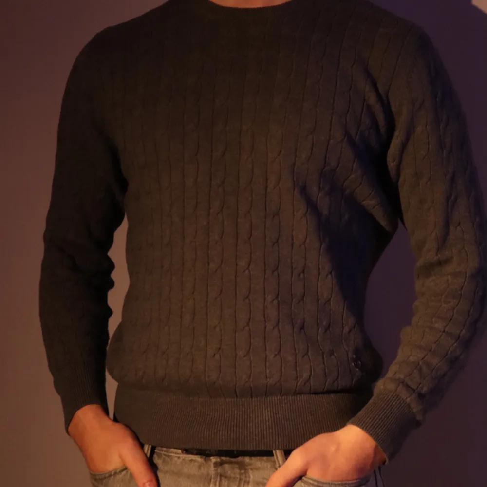 Snygg grå stickad tröja med kabelmönster och rund hals. Perfekt för kyligare dagar med ribbade muddar vid ärmslut och nederkant. En diskret metallknappdetalj ger extra stil. Size: s men passar medium bra. Modellen är 186 cm.. Neuleet.