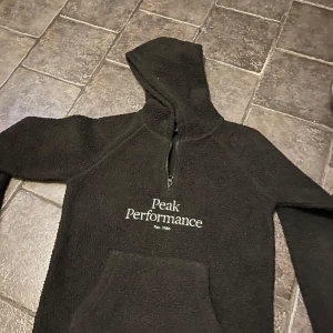 Svart fleece hoodie från Peak Performance - Mysig svart fleece hoodie från Peak Performance med dragkedja och känguruficka. Perfekt för kyliga dagar med sin varma och bekväma design. Märkeslogga på bröstet ger en stilren touch.