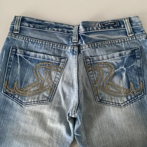 Rock & Republic jeans m. broderade bakfickor  - Snygga blå jeans från Rock & Republic med unika broderade detaljer på bakfickorna. Jeansen har en klassisk femficksdesign och en ljus tvätt med slitna detaljer vid bensluten. Perfekta för en avslappnad stil.