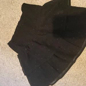 Svart glittrig kjol - Snygg svart kortkjol med glittrande detaljer. Kjolen har en volangdesign och en elastisk midja för bekväm passform. Perfekt för att lägga till lite glamour till din outfit.