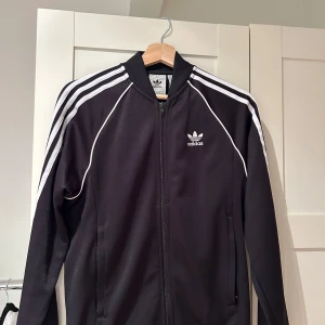 Adidas SST - Säljer en klassisk svart träningsjacka från Adidas med vita ränder längs ärmarna och dragkedja framtill. Jackan har det ikoniska Adidas-loggan på bröstet och är perfekt för sportiga aktiviteter. Aldrig använt fick den i present i en liten storlek 