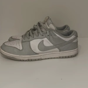 Grå Nike Dunk sneakers - Snygga grå Nike Dunk sneakers med klassisk design och snörning. Perfekta för en stilren look. Skorna har en diskret Nike-logga på sidan och är i en neutral grå färg som passar till det mesta.