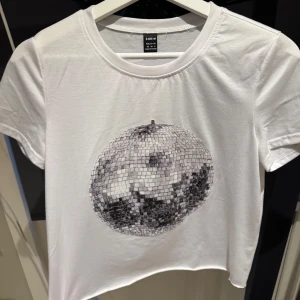 Vit t-shirt med discokulmotiv  - Snygg vit t-shirt från Shein med ett coolt discokulmotiv i silver på framsidan. Perfekt för en avslappnad stil med en touch av festkänsla. Kortärmad och i mjukt bomullsmaterial. Om ni vill ha bilder på hur den sitter på skriv privat
