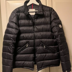Svart dunjacka från Moncler - Snygg svart dunjacka från Moncler med quiltad design och dragkedja framtill. Jackan har långa ärmar och praktiska fickor med dragkedjor. Perfekt för kyligare väder. Storlek L men passar M.