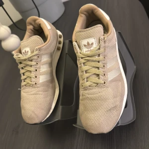 Beige Adidas sneakers - Snygga beige sneakers från Adidas med klassiska tre ränder i vitt på sidan. Skorna har snörning och en bekväm sula med dekorativa detaljer. Perfekta för en avslappnad stil.