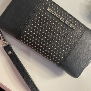 Svart plånbok från Michael Kors - Snygg svart plånbok från Michael Kors med silverfärgade nitar och logotyp. Plånboken har en praktisk dragkedja och en avtagbar handledsrem. Perfekt för att hålla ordning på kort och kontanter. Hör av er av funderingar! 