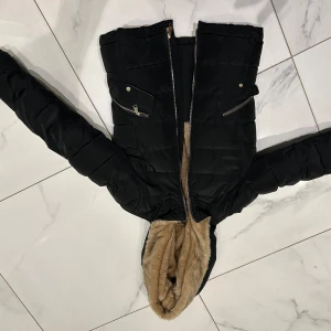 Svart pufferjacka med fuskpäls - Snygg svart pufferjacka med dragkedja och huva kantad med fuskpäls. Jackan har praktiska fickor med dragkedjor och tryckknappar. Perfekt för kyliga dagar.