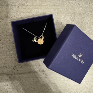 Halsband med stjärntecken från Swarovski - Elegant halsband från Swarovski med ett stjärntecken och en rund berlock. Kedjan är i silverfärg och berlocken har en gnistrande detalj i mitten i rosé guld. Perfekt för att ge en personlig touch till din stil. 