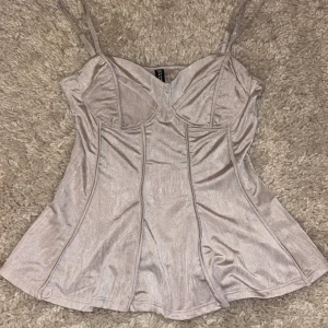 Grå metallic korsettopp med smala axelband - Snygg korsettopp med smala axelband och v-ringad design. Toppen har en figurnära passform med sömmar som framhäver silhuetten. Perfekt för en stilren look. Knappt kommit till användning.