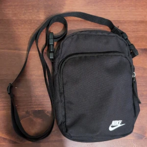 Svart axelväska från Nike - Snygg och praktisk svart axelväska från Nike med justerbar rem och dragkedja. Perfekt för att bära med sig det viktigaste på språng. Diskret Nike-logga i vitt på framsidan.