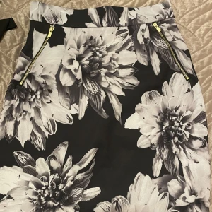 Svart och vit blommig kjol med dragkedja - Snygg svart och vit blommig kjol med dragkedjor fram och bak. Kjolen har ett stilrent blommönster och är perfekt för att ge en elegant touch till din outfit.