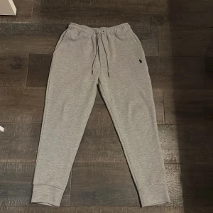 Grå mjukisbyxor från Polo Ralph Lauren - Snygga grå mjukisbyxor från Polo Ralph Lauren med elastisk midja och snörning. Byxorna har en liten broderad logga på sidan och är perfekta för en avslappnad stil. De har även fickor för extra bekvämlighet. Från tracksuit med den gråa zipper
