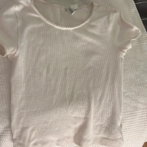 Vit ribbad topp från H&M Divided - Säljer en söt vit ribbad topp från H&M Divided. Toppen har korta ärmar och en fin spetsdetalj vid halsringningen. Perfekt för en avslappnad stil.