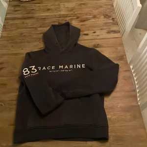 Svart hoodie från Race Marine - Säljer en svart hoodie från Race Marine med tryck på bröstet. Den har en klassisk design med huva och långa ärmar. Perfekt för en avslappnad stil.