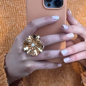 Guldfärgad ring med blomdesign - Stilren guldfärgad ring med en stor blomformad design och en pärla i mitten. Perfekt för att ge en elegant touch till din outfit.