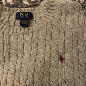 Raulph lauren tröja - Säljer en stilren grå stickad tröja från Ralph Lauren med kabelstickat mönster. Tröjan har långa ärmar och en rund halsringning. Perfekt för både tjejer o killar. Storleken är L(14-16 år) men passar mer en vanlig XS/S