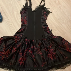 Svart och röd klänning med spetsdetaljer - Snygg svart klänning med röda blommiga detaljer och spets. Klänningen har en dragkedja bak och dekorativa rosetter. Perfekt för en unik stil.