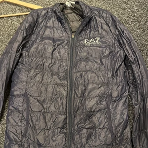 Svart quiltad jacka från EA7 - Snygg svart quiltad jacka från EA7 med dragkedja framtill. Perfekt för kyligare dagar med sin lätta och stilrena design. Jackan har långa ärmar och en diskret logga på bröstet.