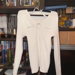 Vit ribbad topp med ringdetalj - Säljer en stilren vit ribbad topp med lång ärm och unik ringdetalj framtill. Toppen har en knytning vid halsen som ger en elegant touch. Perfekt för att lyfta din outfit!