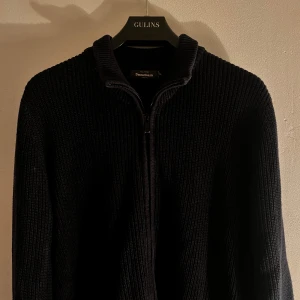 Svart stickad tröja från Dressmann - Snygg svart stickad tröja från Dressmann med dragkedja framtill. Perfekt för kyliga dagar med sin ribbade design och höga krage. .