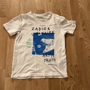 Vit t-shirt från Zadig & Voltaire som är andvänd väldigt få gånger det vill säga 2-3 gånger och är i storlek 146-152 - Snygg vit t-shirt från Zadig & Voltaire med ett coolt blått och svart tryck på framsidan. Perfekt för en avslappnad stil. T-shirten har korta ärmar och rund halsringning.