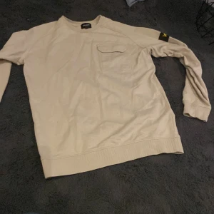 Beige tröja från Lyle & Scott Junior - Säljer en stilren beige tröja från Lyle & Scott Junior. Tröjan har en bröstficka med lock och ett broderat emblem på ärmen. Perfekt för en avslappnad stil.