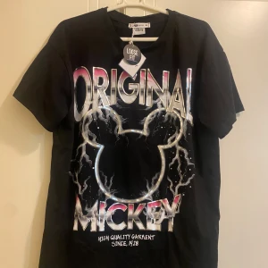Svart Disney T-shirt med Mickey Mouse-tryck - Cool svart T-shirt från Disney med stort 'Original Mickey' tryck i färgglada bokstäver och en silhuett av Mickey Mouse. T-shirten har en loose fit och är perfekt för alla Disney-fans.