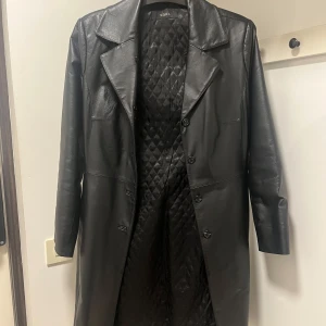 Svart kappa i äkta läder från Saki - Snygg svart skinnkappa från Saki med quiltat foder och avtagbar pälsimitation på kragen. Perfekt för kyligare dagar med sin långa design och knäppning framtill. Kappan är i bra skick men finns tecken på användning. Finns några fläckar, de flesta är inte så synliga (se bifogade bilder). I sin helhet är den i bra skick och mycket bra kvalité. En knapp på jackans krage som är lite lös, knappen används till pälsen. Liten i storlek så skulle säga att den passar 38 bättre.