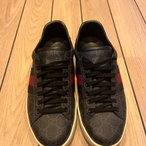 Svarta sneakers med röd detalj från Gucci - Snygga svarta sneakers från Gucci med röd och grön randig detalj på sidan. Skorna har en diskret monogramdesign och svart snörning. Perfekta för en stilren look.