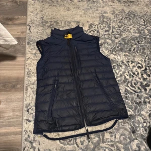 Mörkblå dunväst med dragkedja - Säljer en snygg mörkblå dunväst från Parajumpers. Västen har en dragkedja framtill och en gul detalj vid kragen. Insidan är ljusbeige och västen är perfekt för kyligare dagar. Pris kan diskuteras passar s