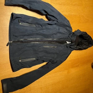 Mörkblå ridjacka med huva - Snygg mörkblå vindjacka från Crw med praktisk huva och dragkedja framtill. Jackan har långa ärmar och flera fickor med dragkedjor för säker förvaring. Perfekt för blåsiga dagar.