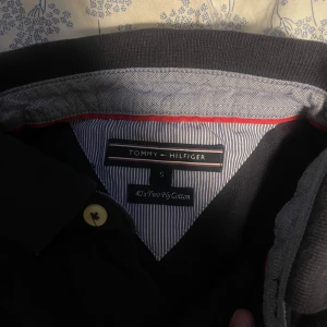 Svart pikétröja från Tommy Hilfiger - Snygg svart pikétröja från Tommy Hilfiger i bomull. Tröjan har en klassisk krage med knappar och det ikoniska Tommy Hilfiger-märket på bröstet. Perfekt för en stilren och avslappnad look.