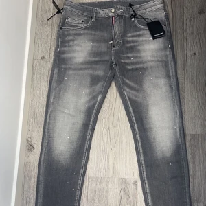Grå Dsquared2 jeans - Snygga grå jeans från Dsquared2 med en cool, sliten look och vita färgstänk. De har en klassisk femficksdesign och knappgylf. Perfekta för en trendig stil. Jeansen är aldrig använda med alla tags kvar, pris kan diskuteras vid snabbaffär 