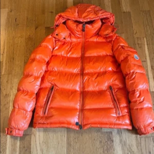 Orange dunjacka från Moncler - Säljer en snygg orange dunjacka från Moncler med huva och dragkedja. Jackan har en quiltad design och praktiska fickor med dragkedjor. Perfekt för kyliga dagar. Tyvär tappat bort kvittot men tags och det funkar. Säljer därav lite billigare👍