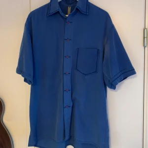 Blå kortärmad skjorta - Snygg blå kortärmad skjorta med röda knappar och en bröstficka. Perfekt för en avslappnad stil.