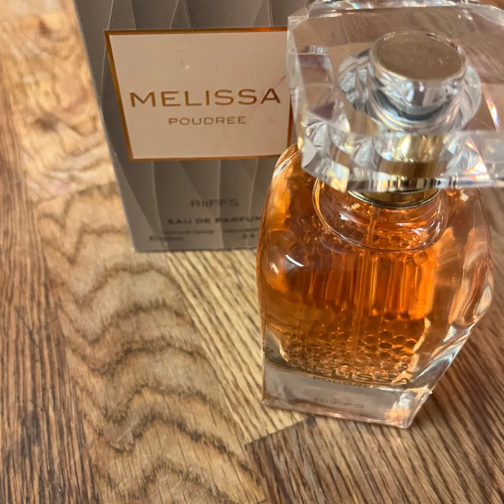 Melissa Poudree Eau de Parfum från Riffs kommer i en elegant glasflaska med en gyllene vätska. Flaskan har en fyrkantig form med en dekorativ, kristalliknande kork. Förpackningen är stilren i grått med guldiga detaljer.. Perfume.