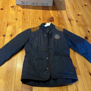 Svart quiltad jacka från Lexington - Snygg svart quiltad jacka från Lexington med bruna axelpartier och broderad logga på bröstet. Jackan har knappar framtill och en stilren design som passar perfekt för höst och vår. Perfekt för dig som gillar en klassisk och tidlös stil.