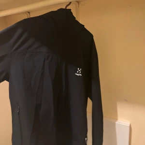 Svart jacka med huva från Haglöfs - Säljer en stilren svart jacka med huva från Haglöfs. Jackan har en dragkedja framtill och är perfekt för utomhusaktiviteter. Den är designad för att ge komfort och skydd i olika väderförhållanden.