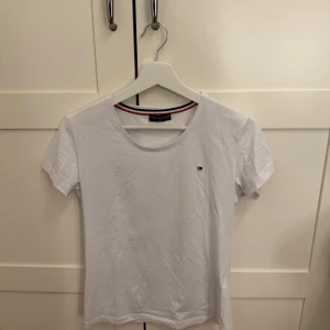 Vit t-shirt från Tommy Hilfiger - Säljer en klassisk vit t-shirt från Tommy Hilfiger med rund hals och korta ärmar. Den har en liten logotyp på bröstet och en stilren design med röd och blå rand på insidan av kragen.