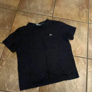 Mörkblå t-shirt från Lacoste - Säljer en klassisk svart t-shirt från Lacoste med den ikoniska krokodilloggan på bröstet. T-shirten har en normal passform och är kortärmad, perfekt för en stilren look. Tröjan har krympt så den motsvarar ungefär storlek 34