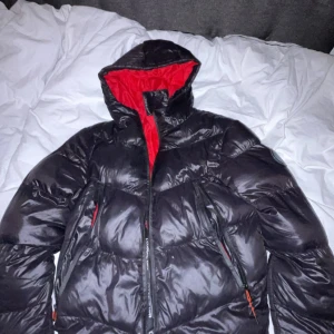 Svart dunjacka från Closure London - Snygg svart dunjacka från Closure London med röd insida. Jackan har en hög krage och dragkedja framtill. Perfekt för kalla dagar med sin pufferdesign. Ett litet hål i vänster arm