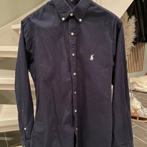 Mörkblå skjorta från Ralph Lauren - Snygg mörkblå skjorta från Ralph Lauren i slim fit. Skjortan har långa ärmar och knappar framtill samt en broderad logotyp på bröstet. Perfekt för en stilren look.