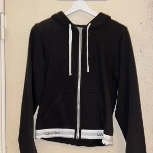 Svart hoodie från Calvin Klein - Säljer en stilren svart hoodie från Calvin Klein med dragkedja och vita snören. Den har en klassisk design med märkets logga på fållen. Perfekt för en avslappnad look.